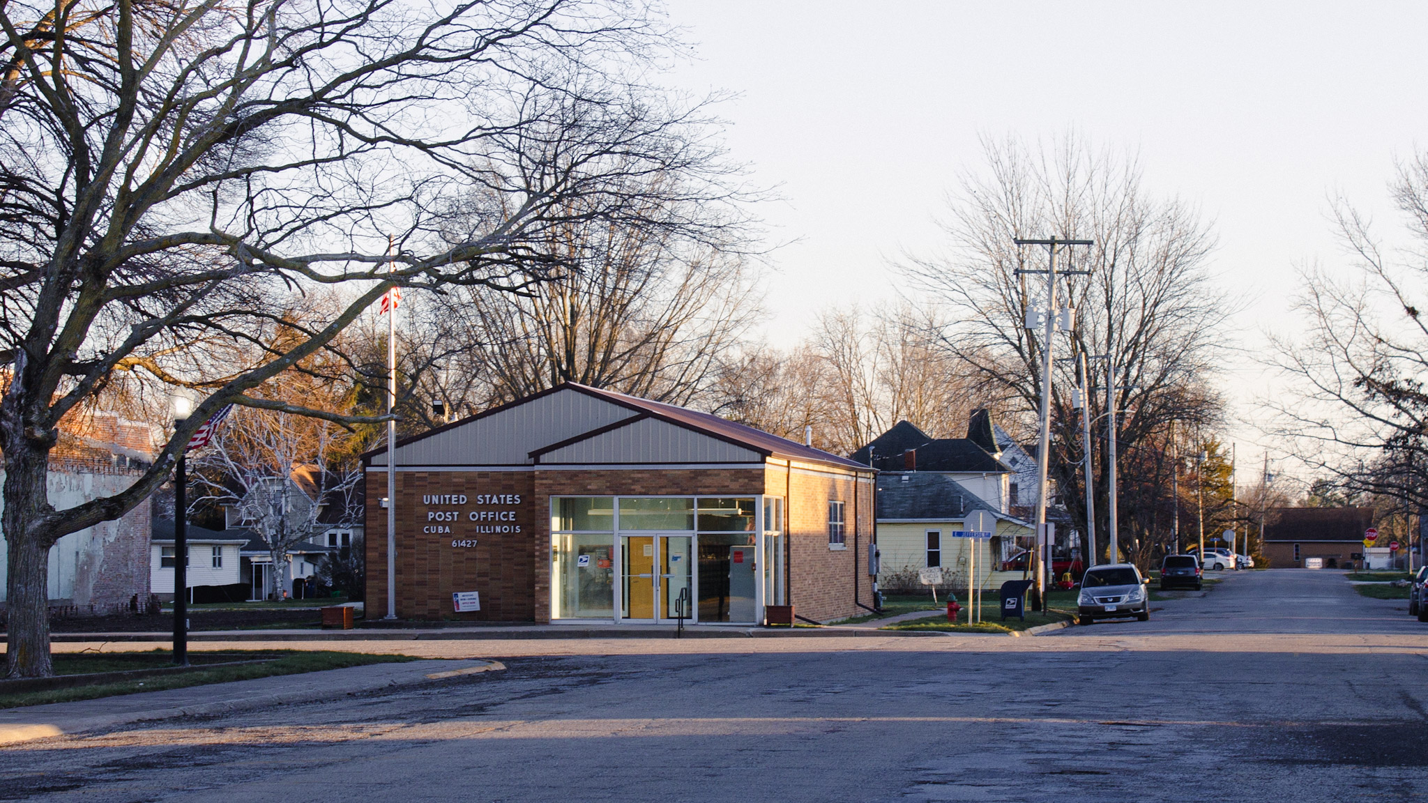 Long shadows fall across an empty street. The bare limbs of a tree stretch over a one-story post office.